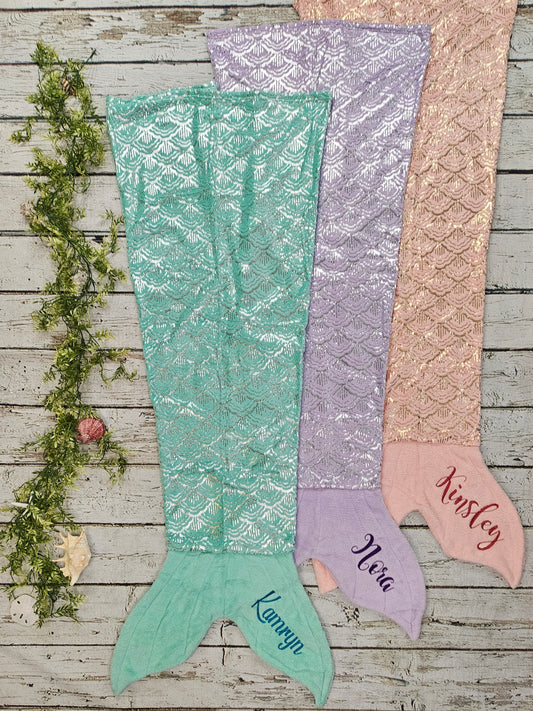 Three mermaid tail blankets in green, purple, and pink on a wooden surface with names embroidered.